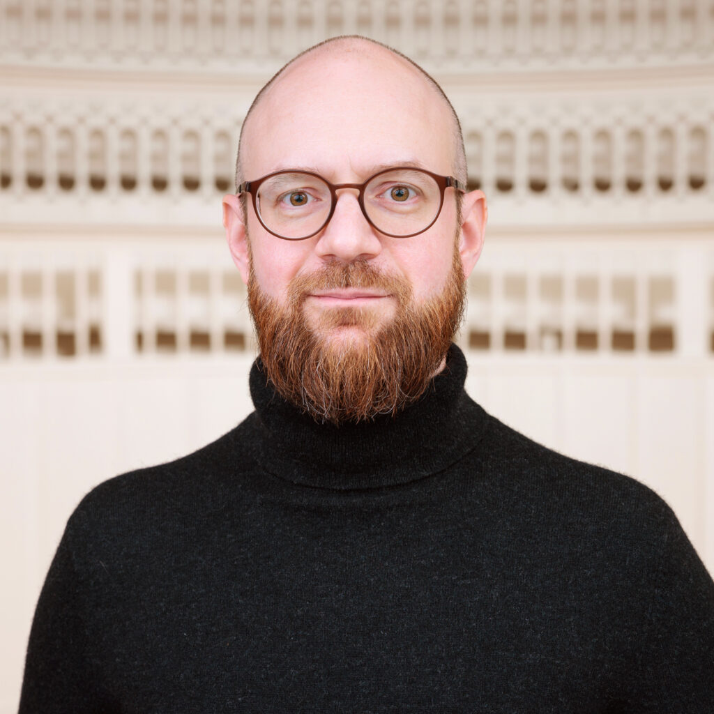 Bust portrait of Felix Sattler by Philip Plum. He is a middle-aged white mal with bald head and a beard, wearing a black turtleneck. He is standing in the Tieranatomisches Theater auditorium.