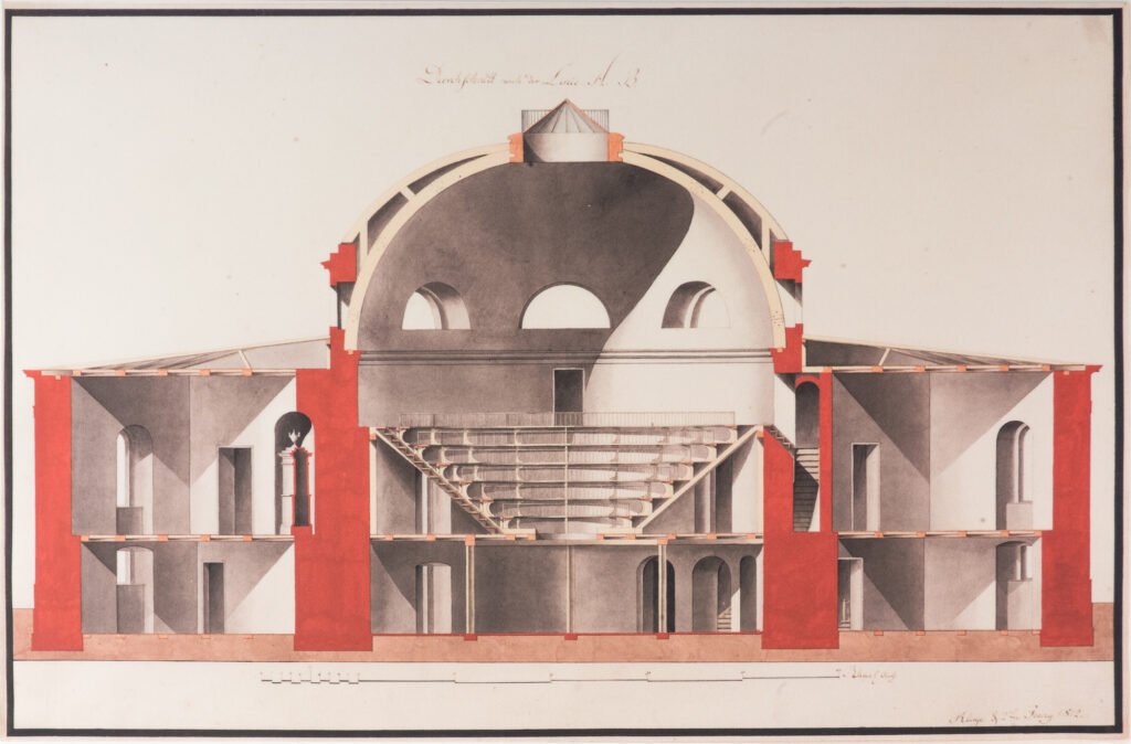 Bild / Architektonische Zeichnung. Lavierte Federzeichnung von Johann Friedrich Kluge aus dem Jahr 1812 mit einem Querschnitt des Tieranatomischen Theaters entlang der Linie AB. Die Zeichnung zeigt den symmetrischen Aufbau des Gebäudes mit dem zentralen, von einer Kuppel überwölbten Hörsaal und den umlaufenden, gestuften Sitzreihen. Beidseitig schließen sich Räume und Nebenbereiche an. Die Schnittdarstellung macht die innere Raumstruktur, Treppen, Türen, Fenster und die Konstruktion der Kuppel sichtbar. Rote Flächen markieren Teile der Gebäudekonstruktion.