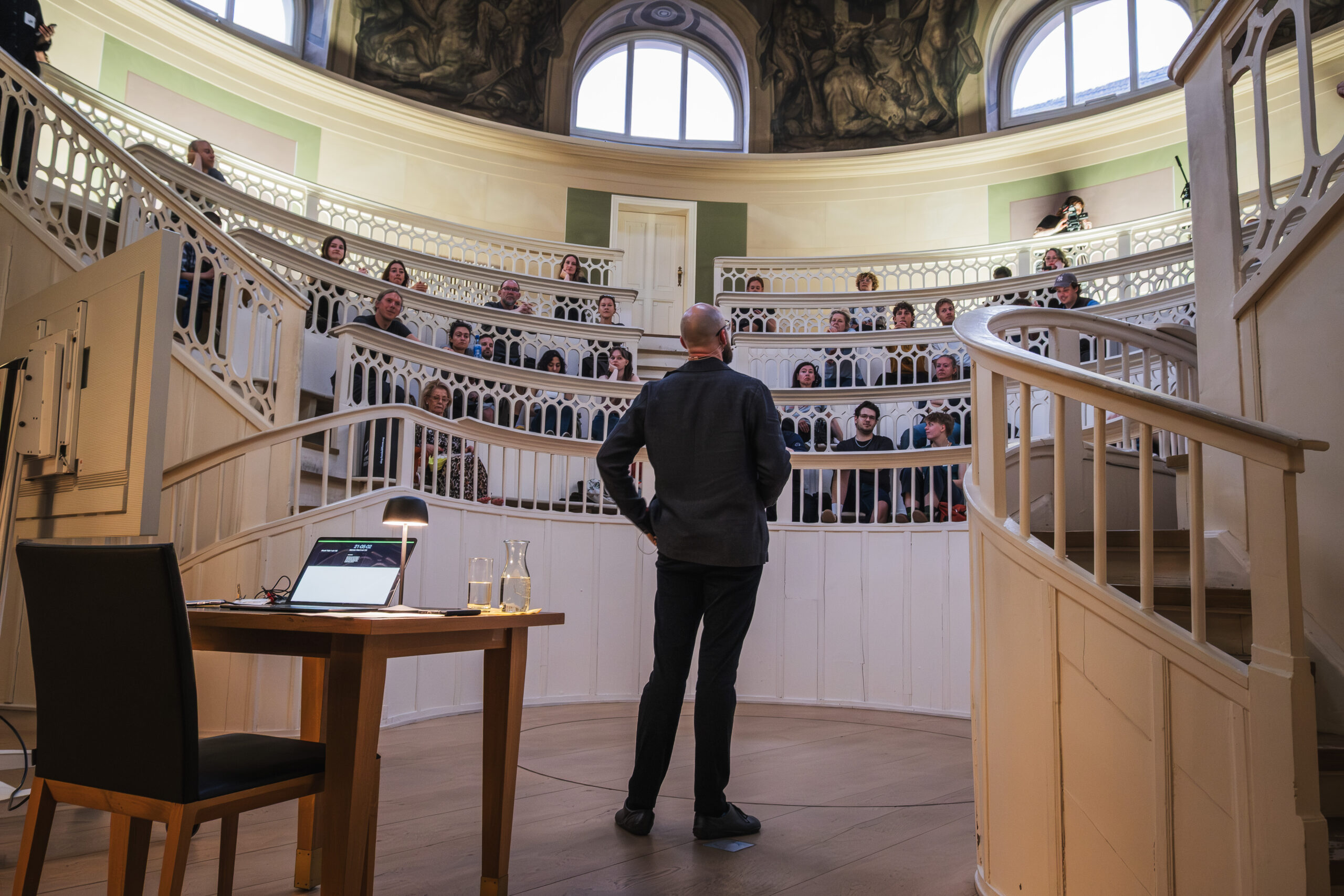 Felix Sattler steht im Zentrum des Tieranatomischen Theaters und spricht zum Publikum. Die Zuhörenden blicken von den umlaufenden Rängen auf die offene Mitte des Raums.