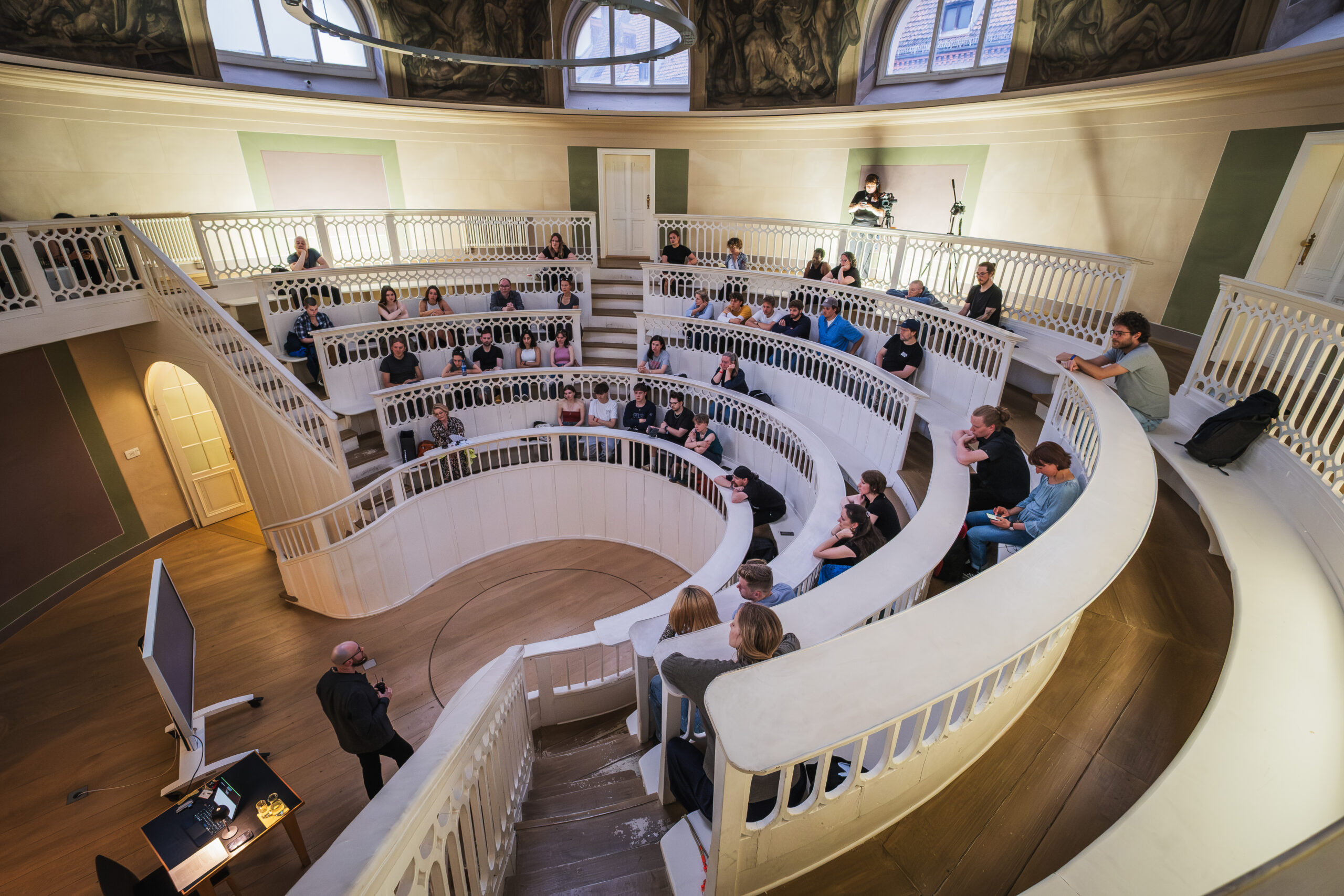 Im kreisförmigen Hörsaal des Tieranatomischen Theaters sitzen rund vierzig Menschen auf geschwungenen weißen Rängen. Im Zentrum begleitet Felix Sattler das Publikum durch eine gemeinsame Hörreise in die Tiefsee.