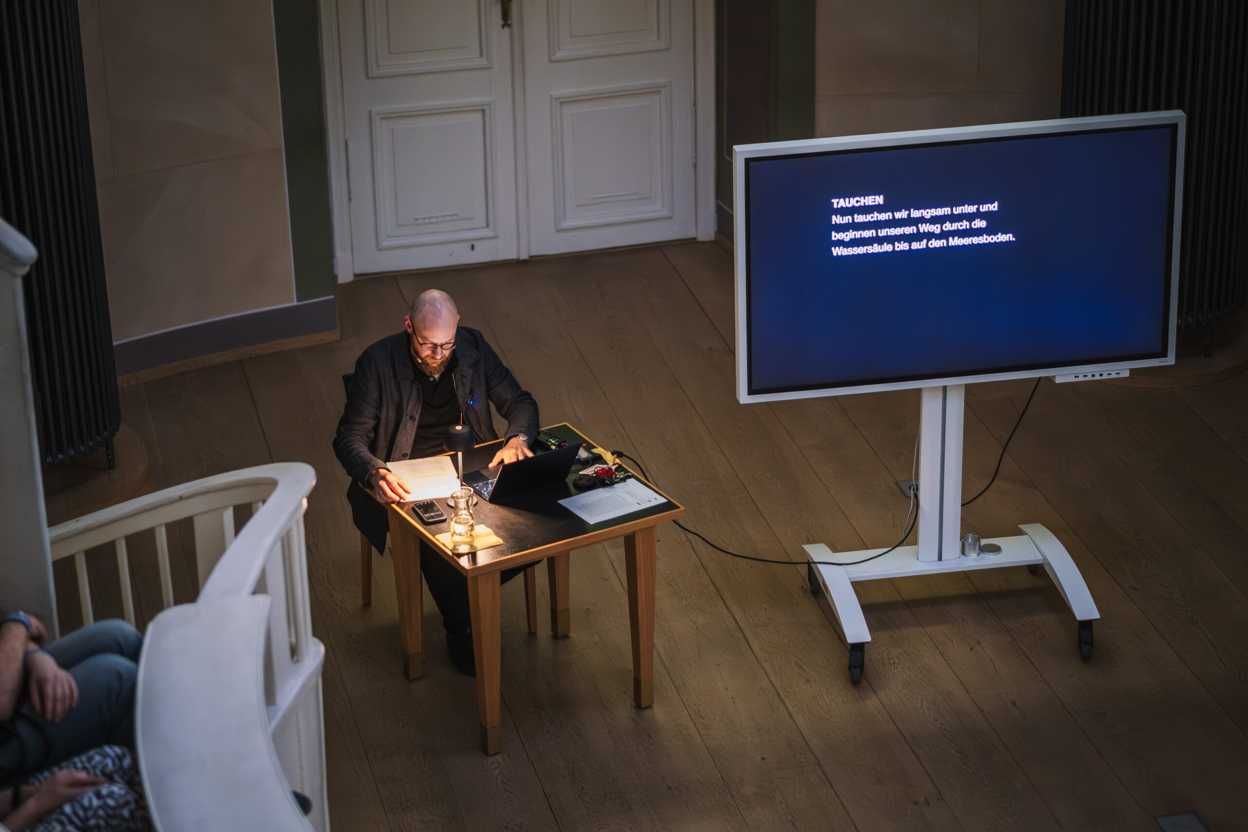 Felix Sattler sitzt an einem kleinen Tisch im Zentrum des historischen Hörsaals im Tieranatomischen Theater. Auf einem Bildschirm erscheint die Einladung, gedanklich in die Tiefsee hinabzutauchen.