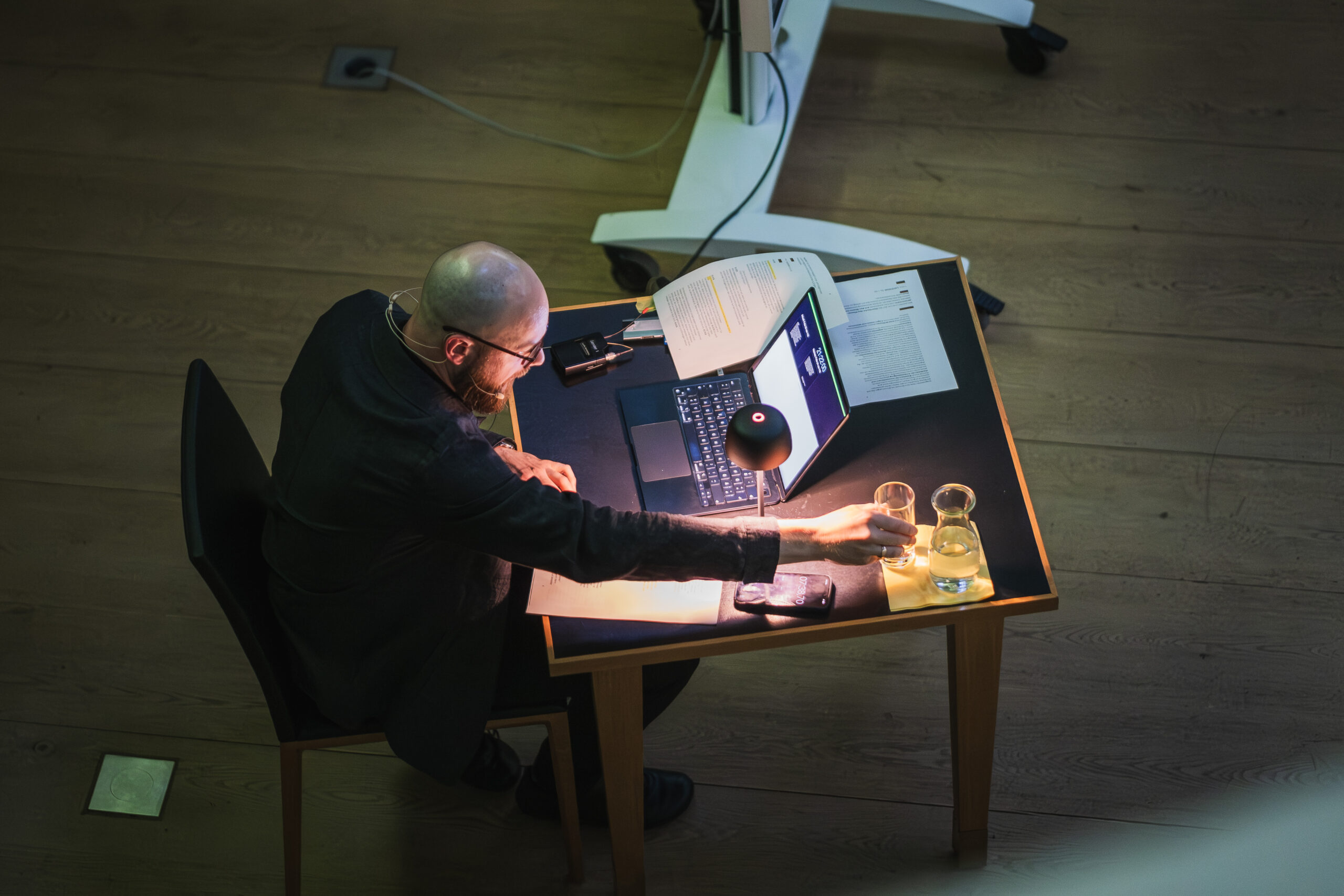 Blick von oben auf Felix Sattler während der Lesung "In die Tiefe spüren" zur Langen Nacht der Wissenschaft 2026 am 6.6.2026. Manuskript, Laptop, Wasserglas und Lampe liegen auf dem Tisch.