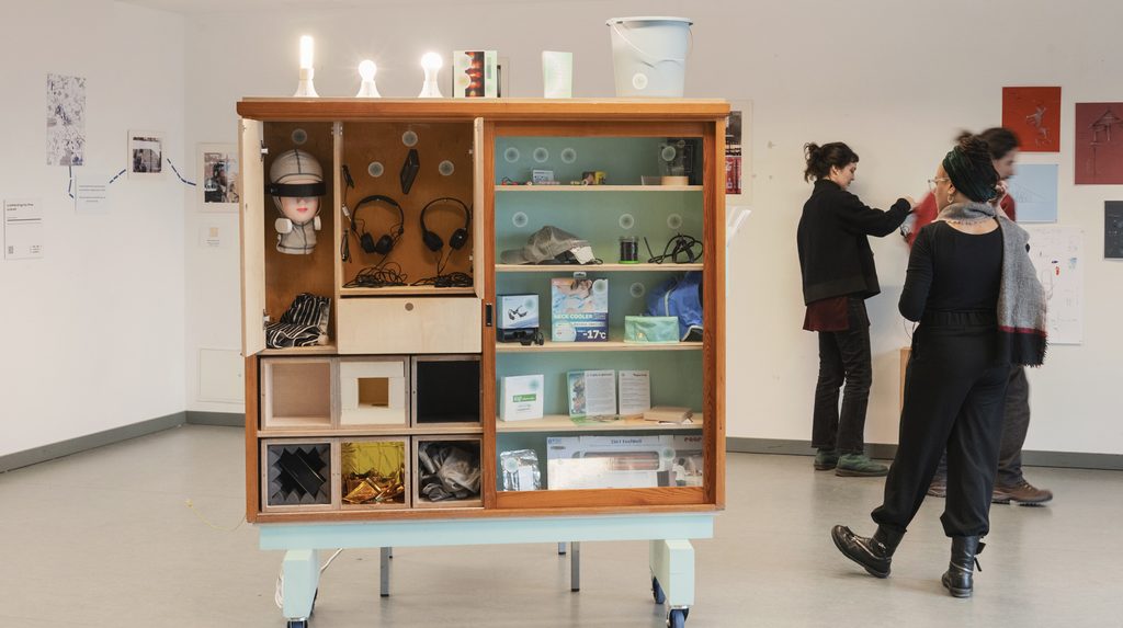 The Cabinet of Waves, a central installation in the exhibition Wavematters, stands on a light-blue wheeled base in an exhibition space. The large open wooden cabinet contains numerous compartments and glass-fronted shelves displaying objects collected as part of the research for Wavematters, including headphones, a mannequin head wearing headphones and an eye covering, packaging for technical devices, fabrics, reflective materials and other everyday objects. Several illuminated light bulbs and a white container are placed on top of the cabinet. Photographs, texts and drawings from the exhibition are displayed on the walls around it. Two people are visible walking through the exhibition space in the background. The Cabinet of Waves brings together objects that explore different experiences and forms of waves in urban environments.