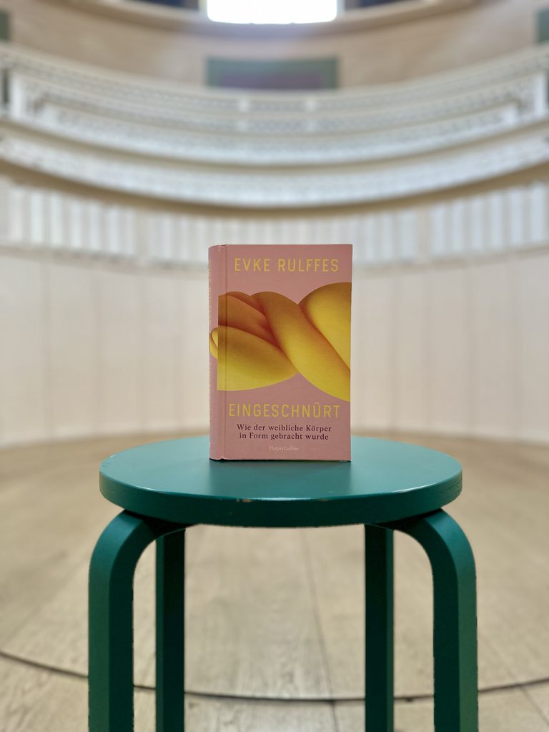 Ein Exemplar von Evke Rulffes’ Buch Eingeschnürt. Wie der weibliche Körper in Form gebracht wurde steht auf einem runden, türkisfarbenen Tisch im historischen Hörsaal des TA T – Tieranatomischen Theaters. Im Hintergrund sind unscharf die geschwungenen, weißen Sitzreihen und Brüstungen des Hörsaals zu erkennen. Das rosafarbene Buchcover zeigt gelbe und orangefarbene, ineinander verschlungene Formen. Die Aufnahme ist frontal komponiert; Buch und Tisch bilden den Mittelpunkt des Bildes.